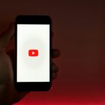 A hand holding a smartphone displaying the YouTube app against a red background.